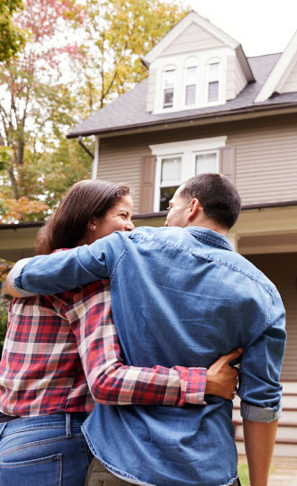 A man and woman smiling with their arms around each other in front of a house.