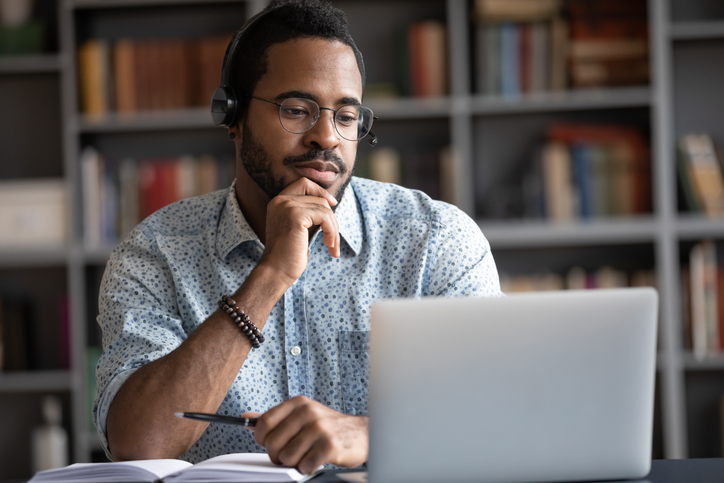 A man wearing head phones with a pen in his hand looking at his laptop.