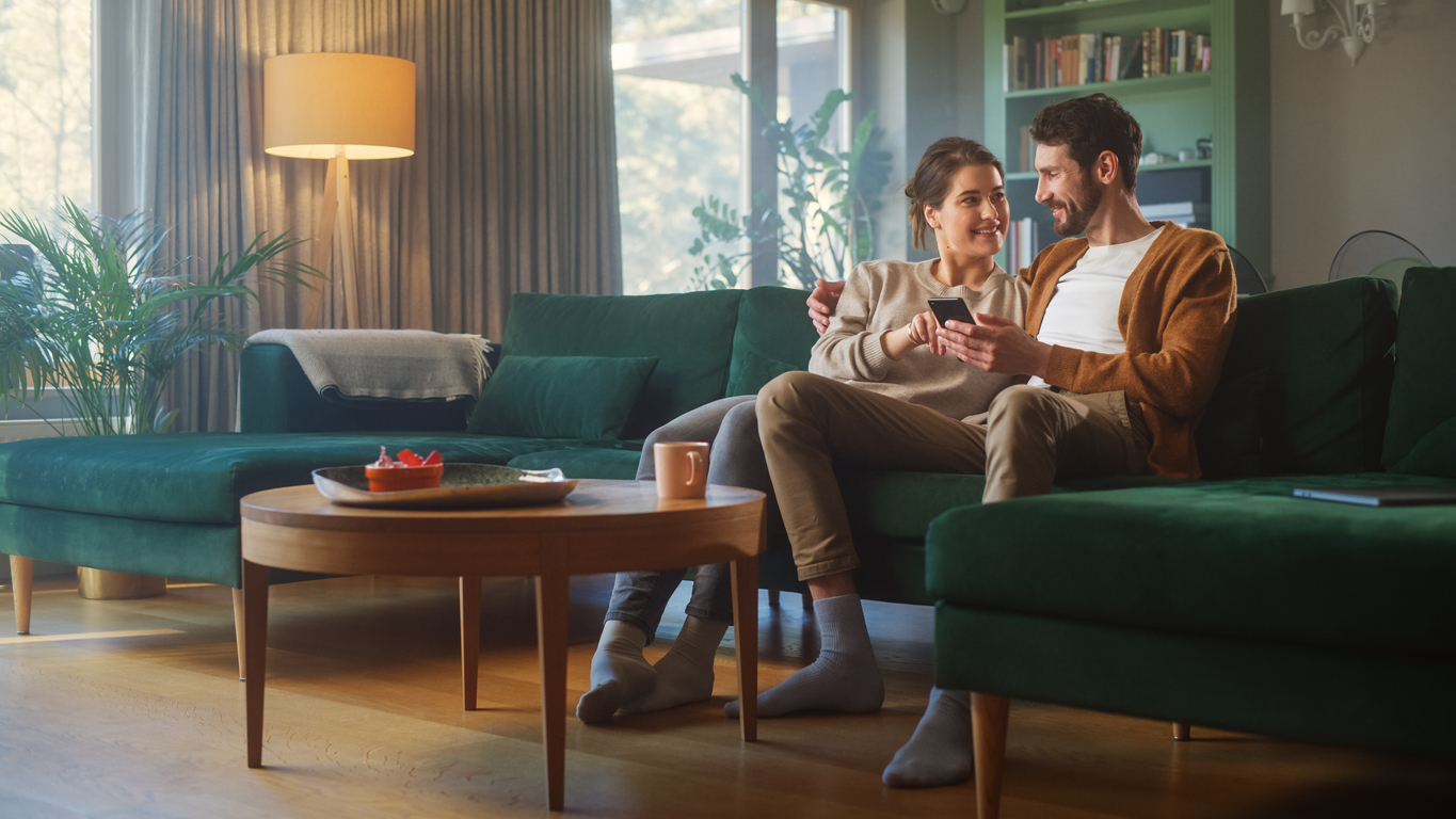 A couple sitting on the couch looking at each other smiling holding a phone.